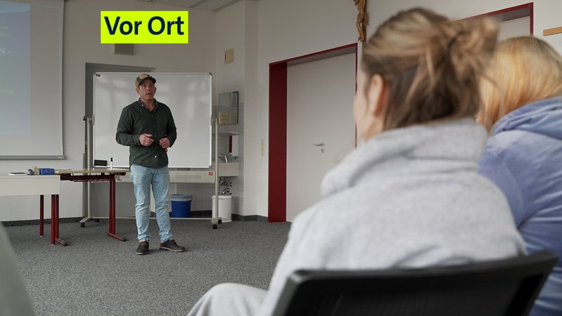Ausbildungsakquisiteur Florian Wagner steht vor einer Klasse. Er ist vorne an der Tafel zu sehen, im Vordergrund sieht man zwei Schülerinnen von hinten. | Bild: BR/Karl Spannenberger Ausbildungsakquisiteur Florian Wagner steht vor einer Klasse. Er ist vorne an der Tafel zu sehen, im Vordergrund sieht man zwei Schülerinnen von hinten.