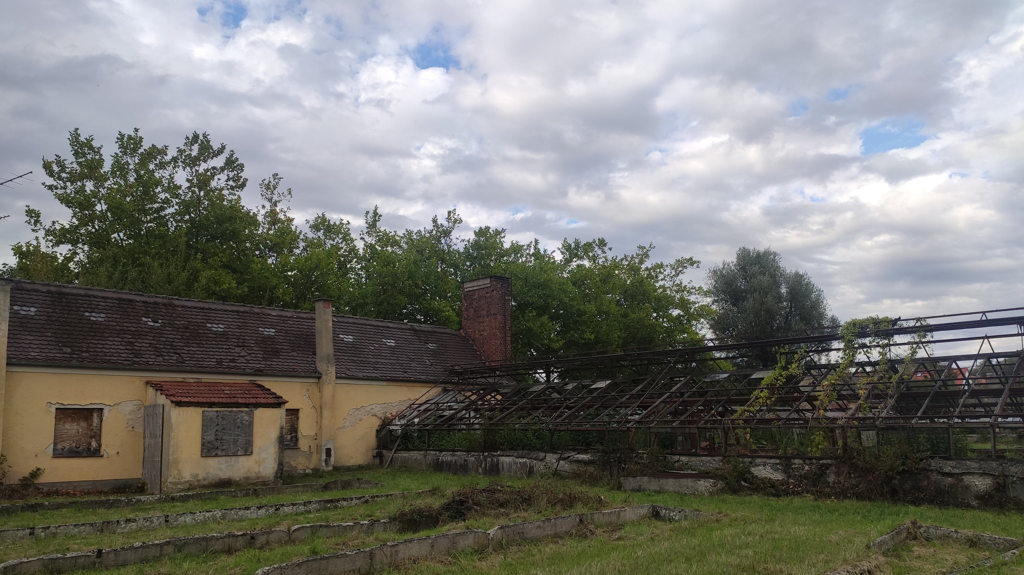 Die Überreste eines Gewächshauses auf der "Plantage" Dachau, daneben ein gelb gestrichenes SS-Verwaltungsgebäude. | Bild: BR/Lisa Westhäußer Die Überreste eines Gewächshauses auf der "Plantage" Dachau, daneben ein gelb gestrichenes SS-Verwaltungsgebäude.