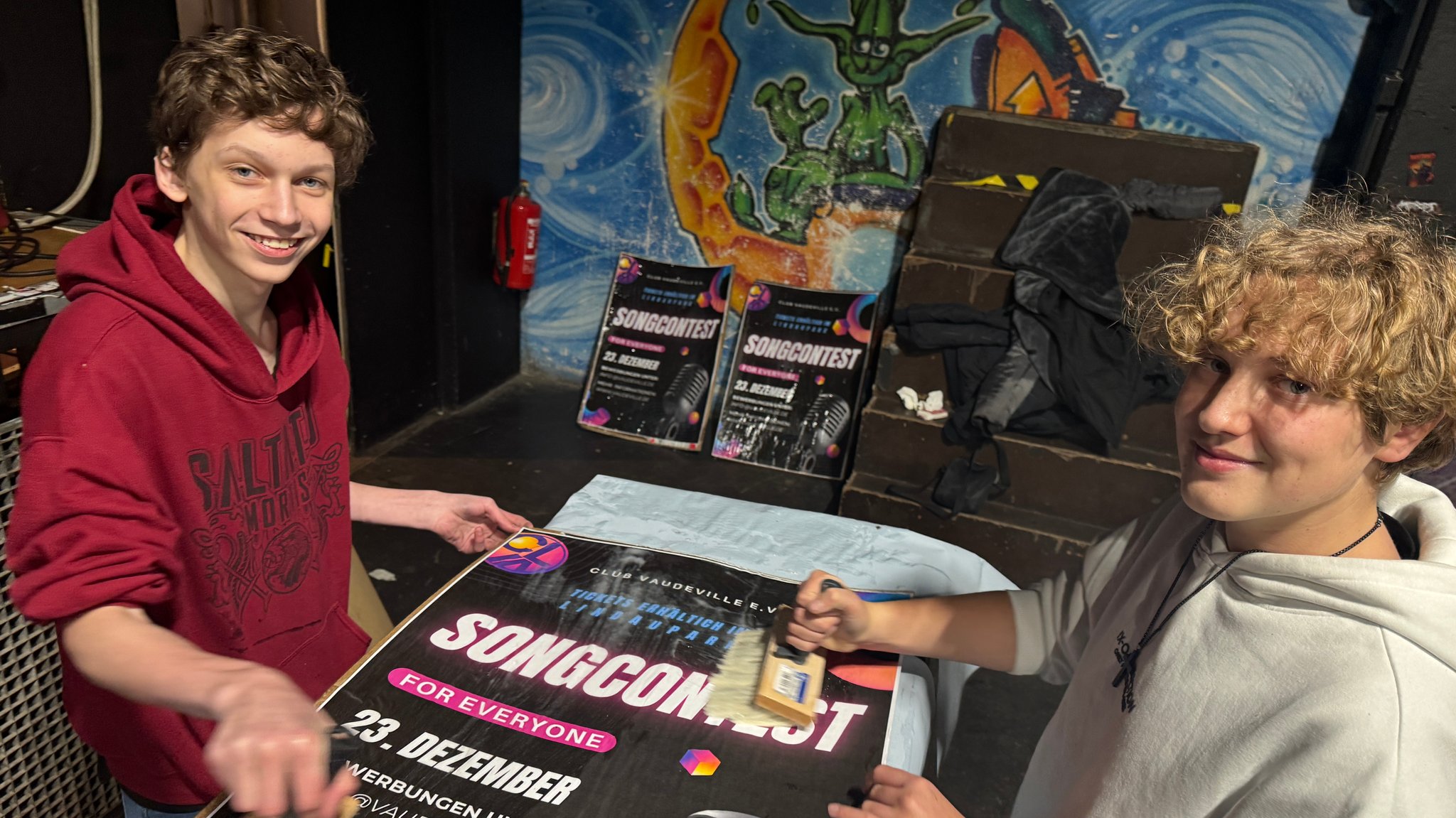 Thorben Schelle (li.) und Luca Jakobsmeier (re.) streichen mit einem breiten Pinsel Kleister auf ein Werbeplakat.