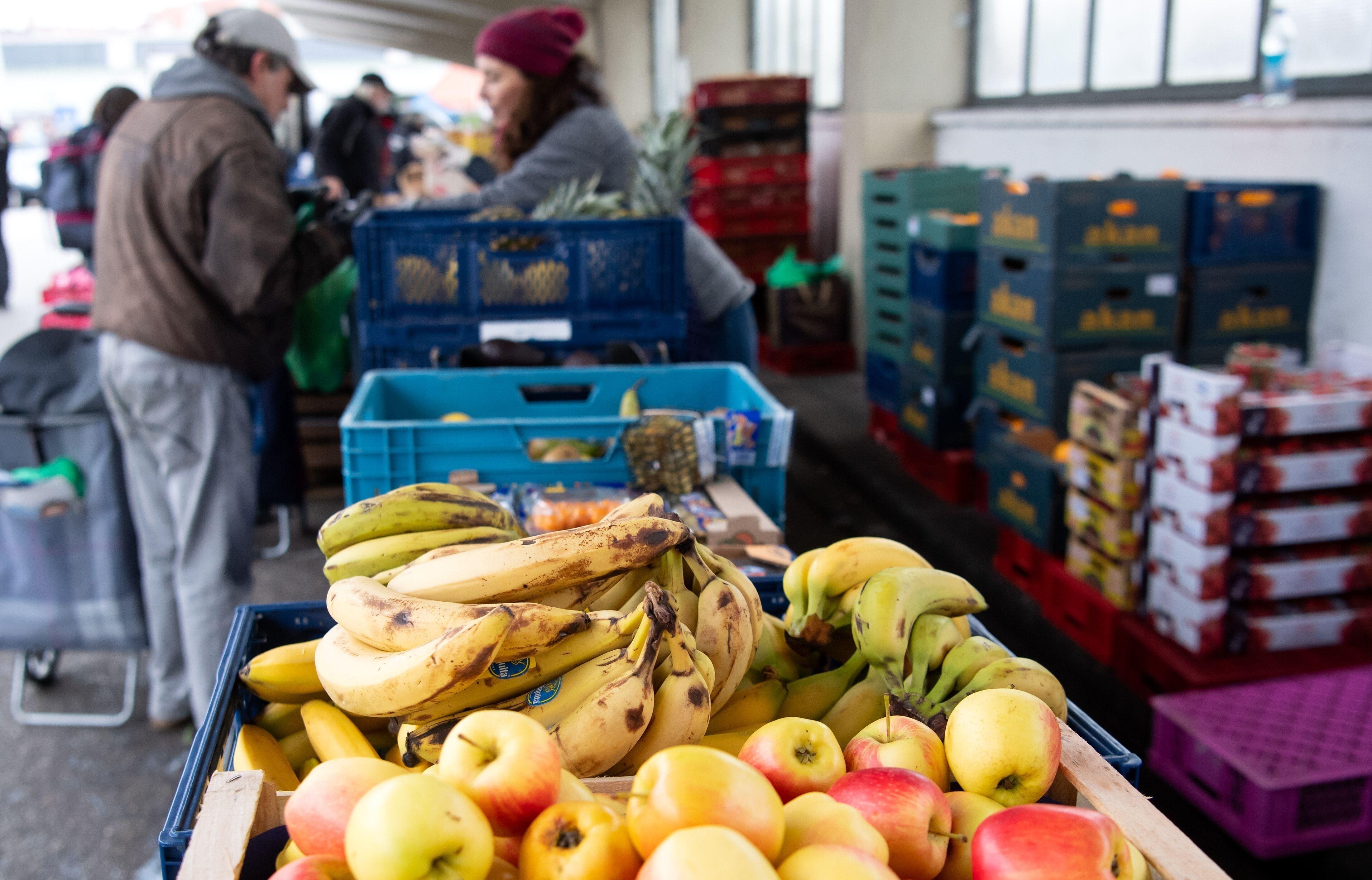 Im Vordergrund liegt Obst, während im Hintergrund Helfende einer Tafel Essen austeilen.