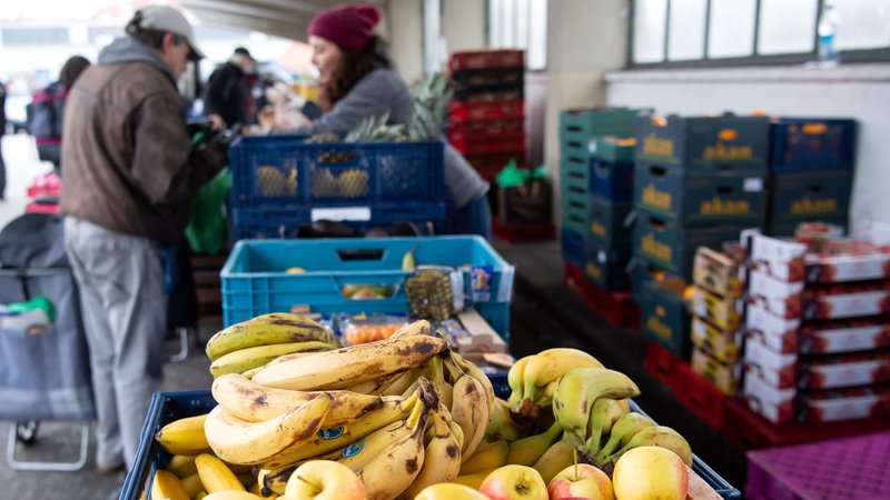Im Vordergrund liegt Obst, während im Hintergrund Helfende einer Tafel Essen austeilen. | Bild: pa/dpa/Sven Hoppe Im Vordergrund liegt Obst, während im Hintergrund Helfende einer Tafel Essen austeilen.
