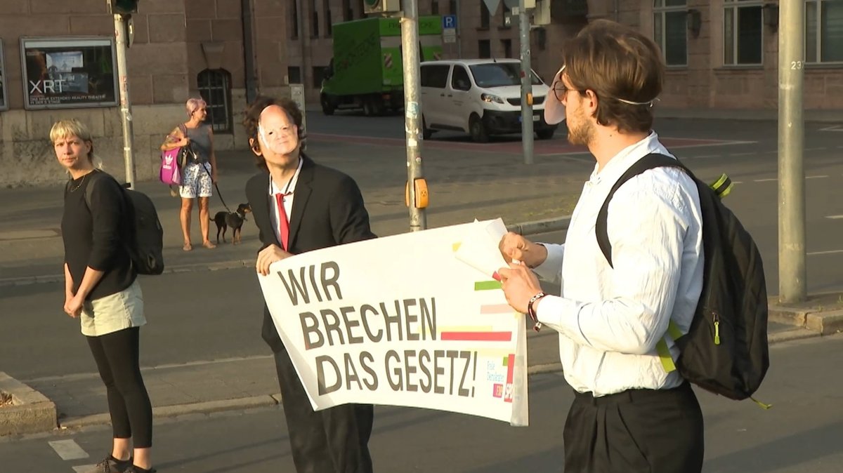 Zwei Klimaaktivisten mit Politikermaske sitzen auf einer Straße und halten ein Transparent mit der Aufschrift "Wir brechen das Gesetz".