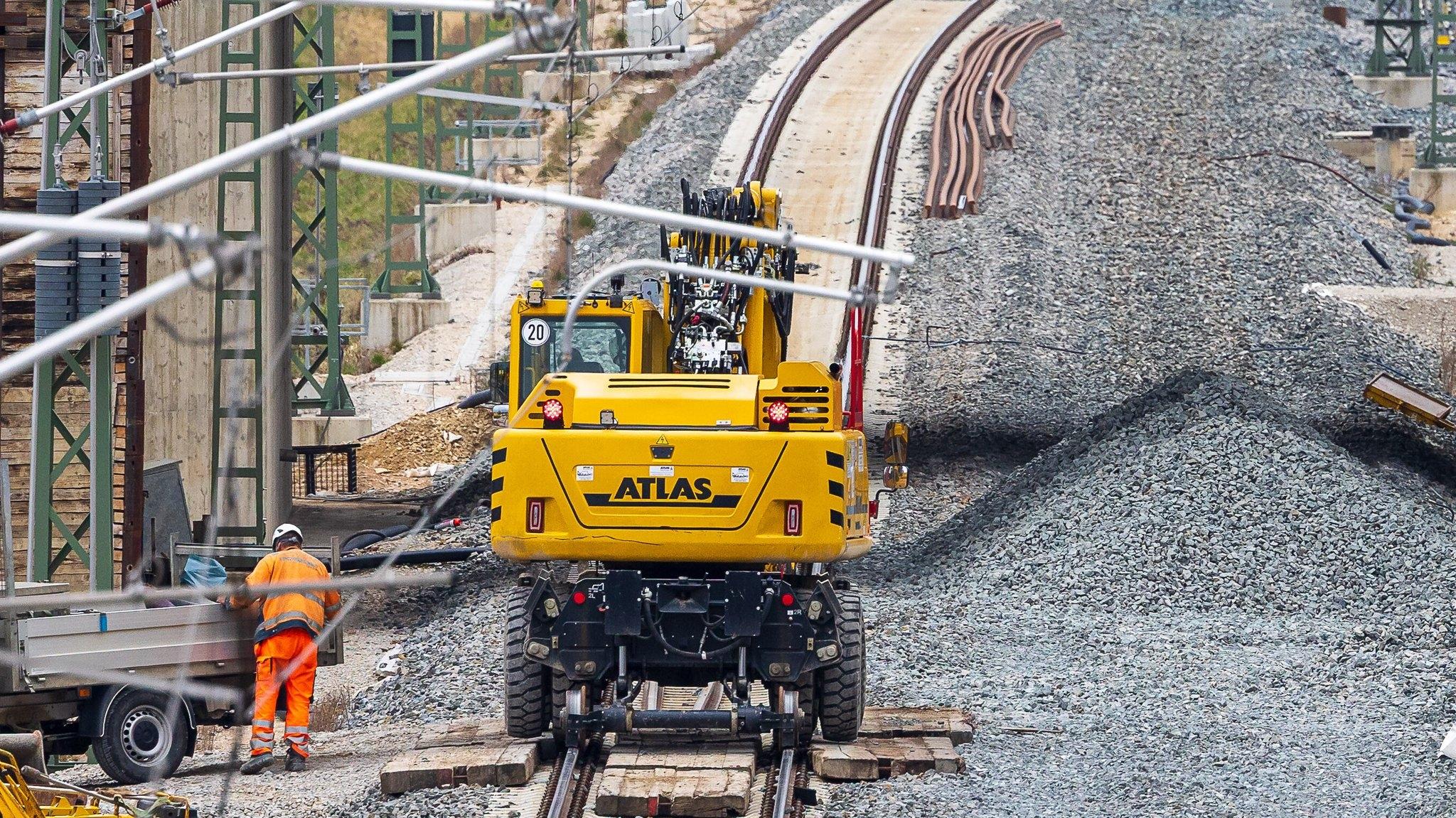 Ein gelber Bagger steht auf einer Zugstrecke, auf der Schienen verlegt werden. 