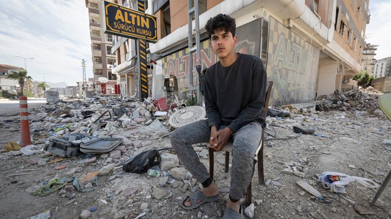 27.4.2023 Antakya: Yusuf sitzt vor einer zerstörten Häuserzeile und beobachtet, wie auf der anderen Straßenseite das Haus seiner Familie abgerissen wird. 