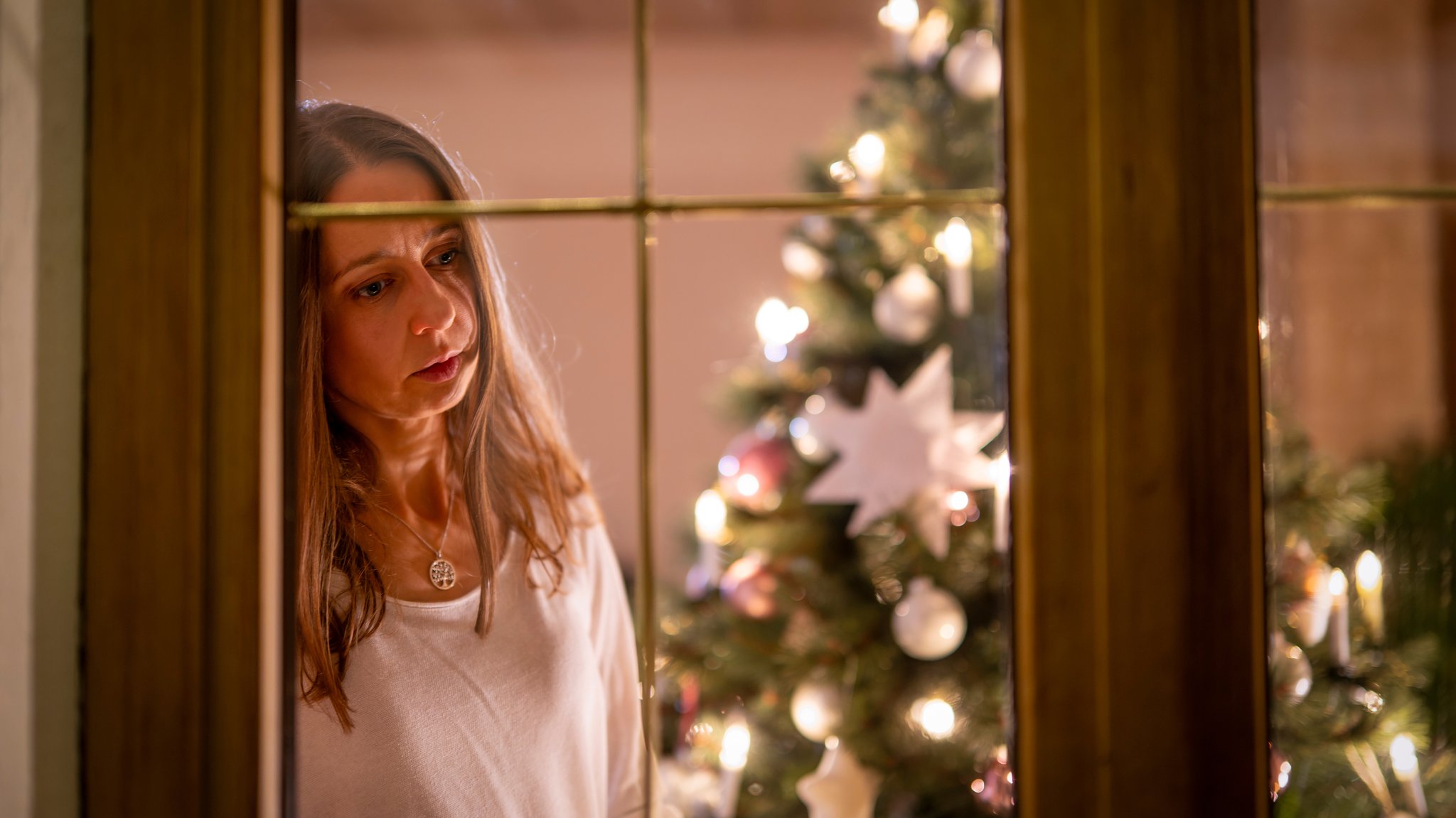 Ein festlich dekorierter Weihnachtsbaum in einer Wohnung im Unterallgäu. Eine Frau schaut nachdenklich aus dem Wohnzimmer durchs Fenster, sie ist alleine. 