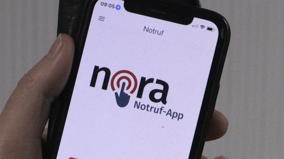 Eine Hand hält ein Handy. Auf dem Display wird die Nora-Notruf-App angezeigt. 