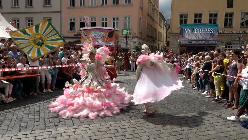 Zwei Tänzerinnen in rosa-weißen Kostümen tanzen auf der Straße, hinter dem Absperrband viel Publikum.