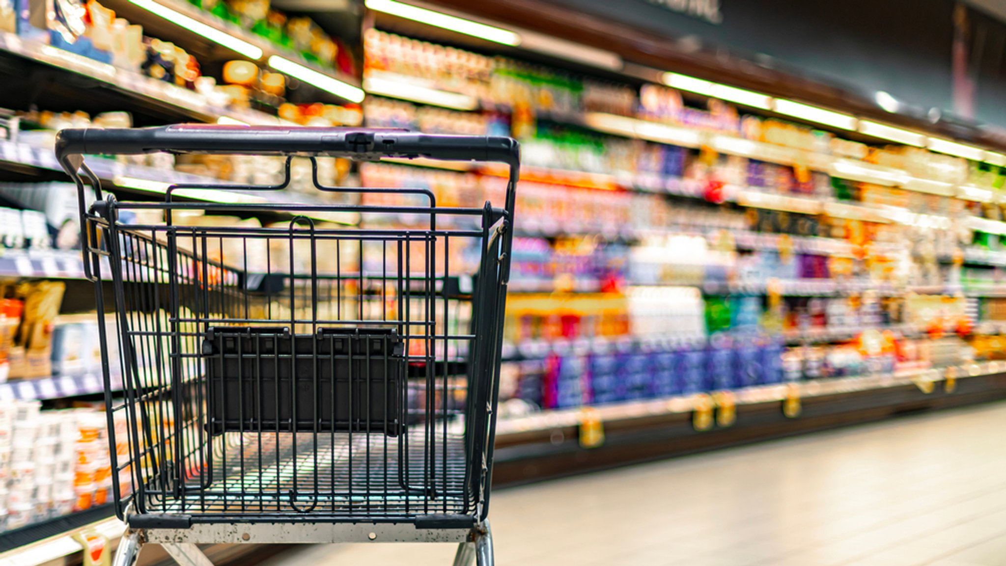 Ein leerer Einkaufswagen steht vor vollen Supermarktregalen. | Bild: stock.adobe.com/monticellllo Ein leerer Einkaufswagen steht vor vollen Supermarktregalen.