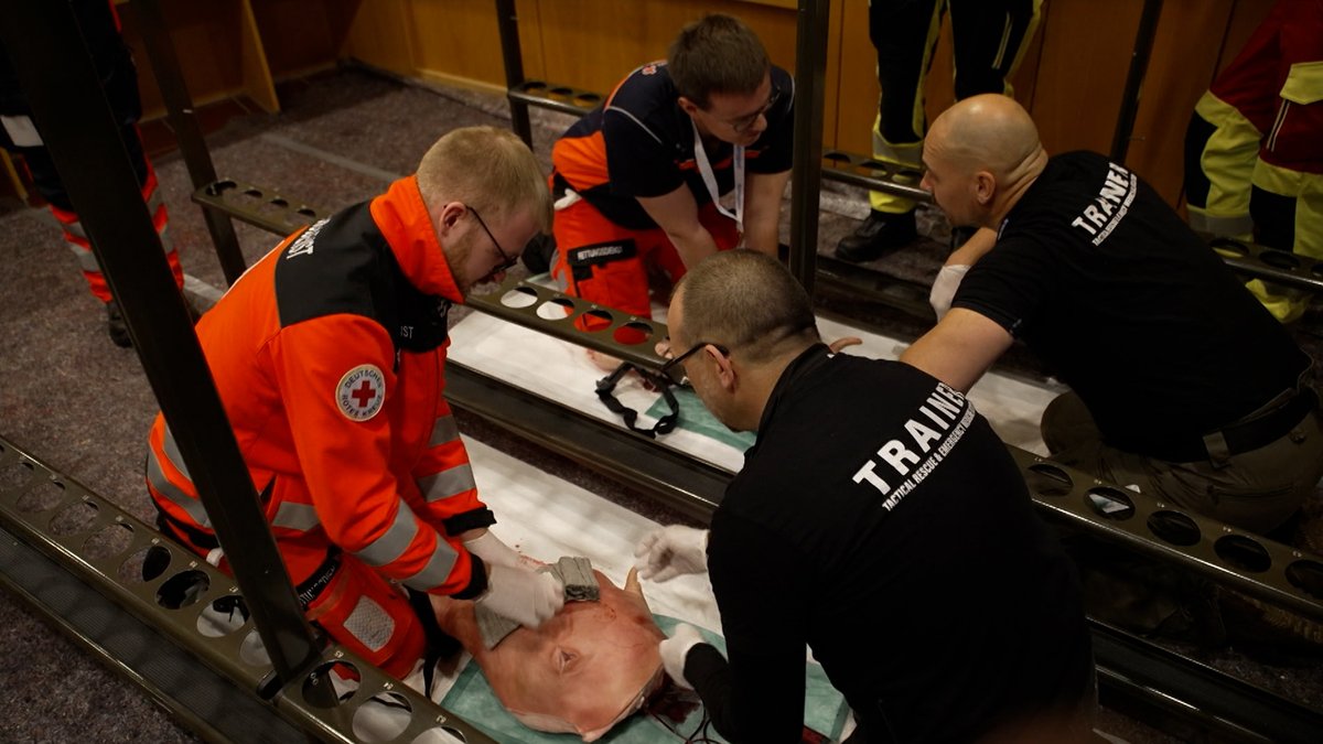 Ein Sanitäter in oranger Warnweste kniet vor dem Hinterschenkel eines Schweins auf dem Boden zwischen engen Garderobenreihen und übt die Versorgung. 