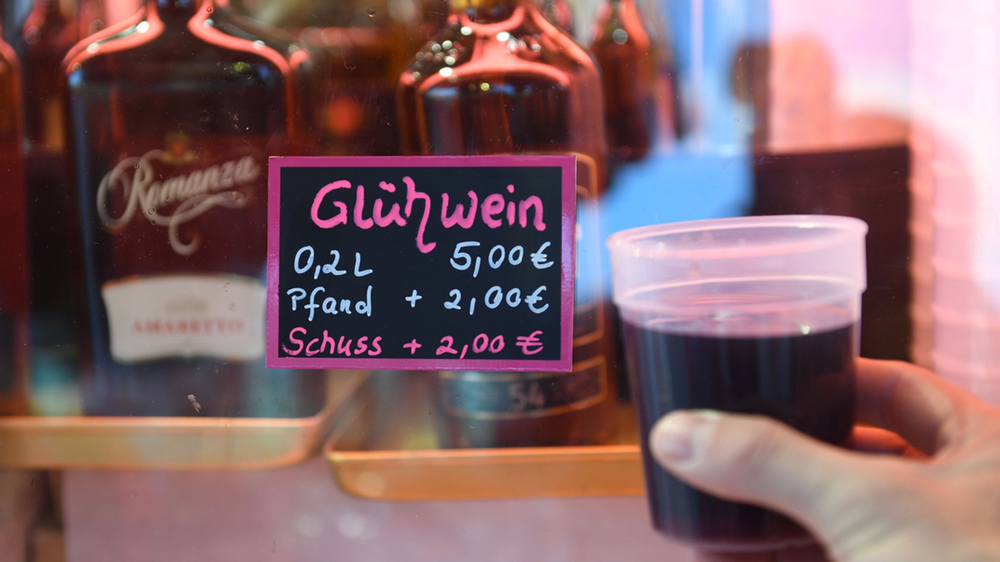 Ein Preisschild weist darauf hin, dass der Glühwein auf der Wiesn fünf Euro plus zwei Euro Pfand kostet. Im Vordergrund ist ein voller Becher mit Glühwein zu sehen.