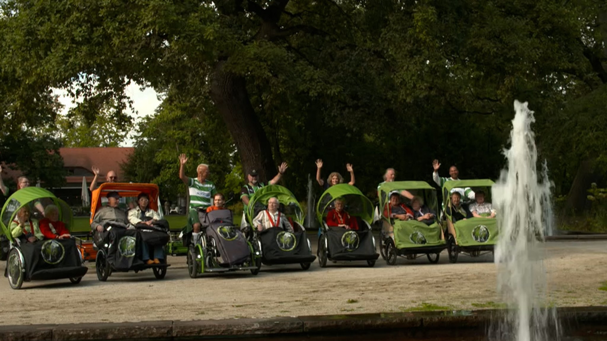 Mehrere grüne Rikschas mit Fahrern und Passagieren stehen in einem Park in einer Reihe.