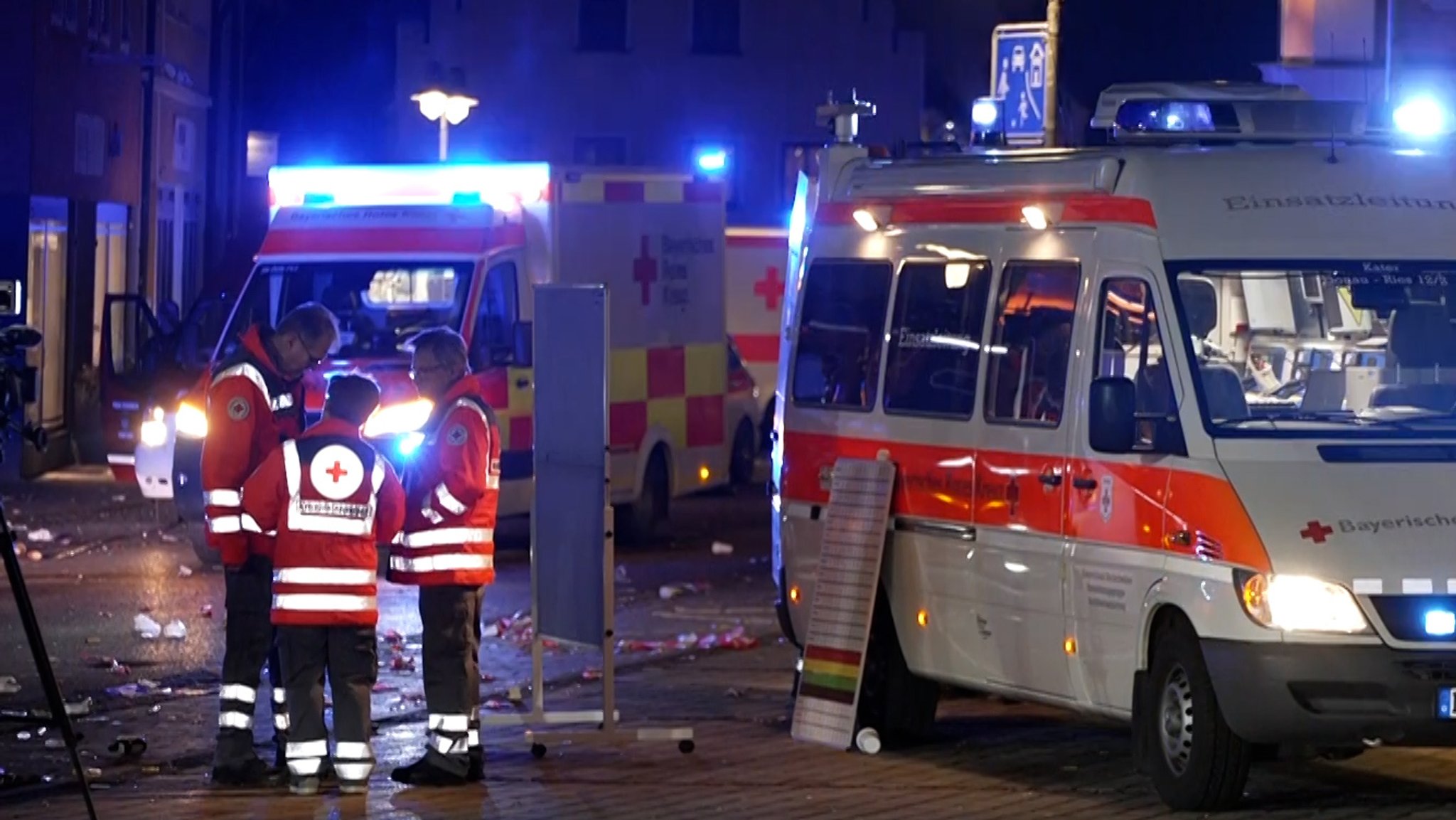 Im Dunklen stehen drei Rettungskräfte vom Bayerischen Roten Kreuz in Einsatzkleidung beieinander auf einer gepflasterten Fläche. Um sie herum stehen mehrere Einsatzwagen.