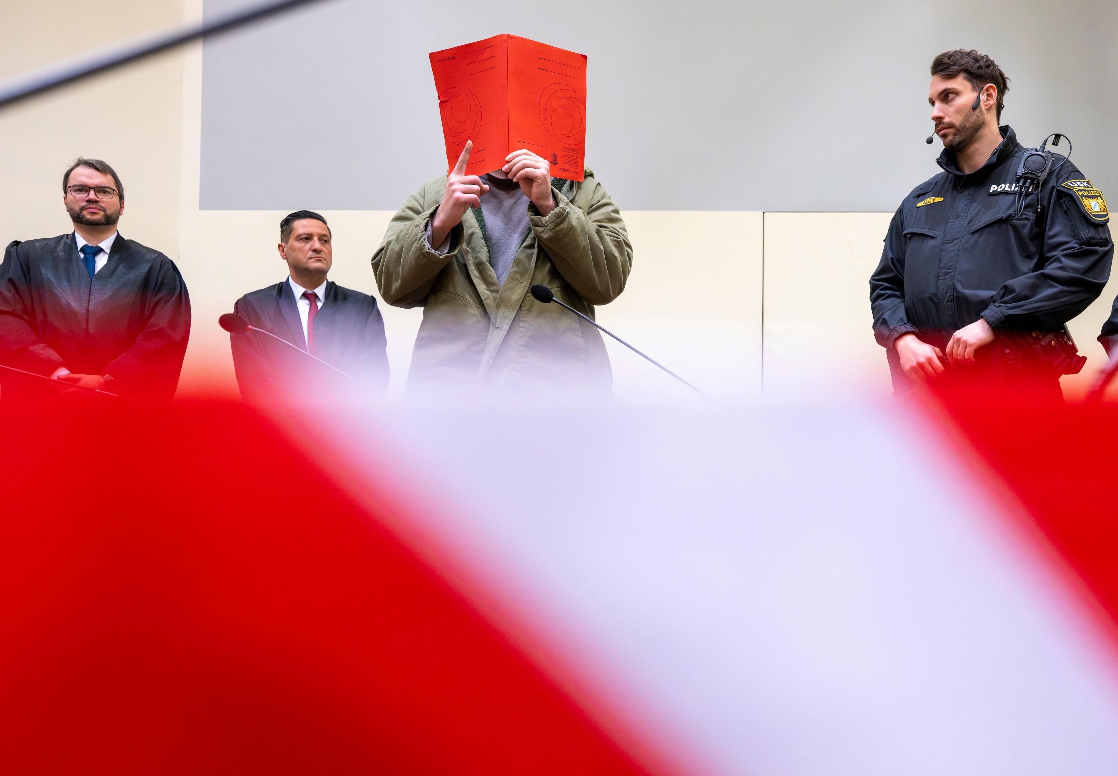 dpatopbilder - 16.01.2026, Bayern, München: Der Angeklagte Farhad N. steht zu Prozessbeginn mit erhobenem rechten Zeigefinger zwischen seinen Anwälten Johann Bund (l) und Ömer Sahinci (2.v.l.) und Polizeibeamten im Prozesssaal an seinem Platz. Farhad N. ist nach einem Auto-Anschlag am 13. Februar 2025 auf eine Demonstration in München des zweifachen Mordes angeklagt. Foto: Peter Kneffel/dpa +++ dpa-Bildfunk +++