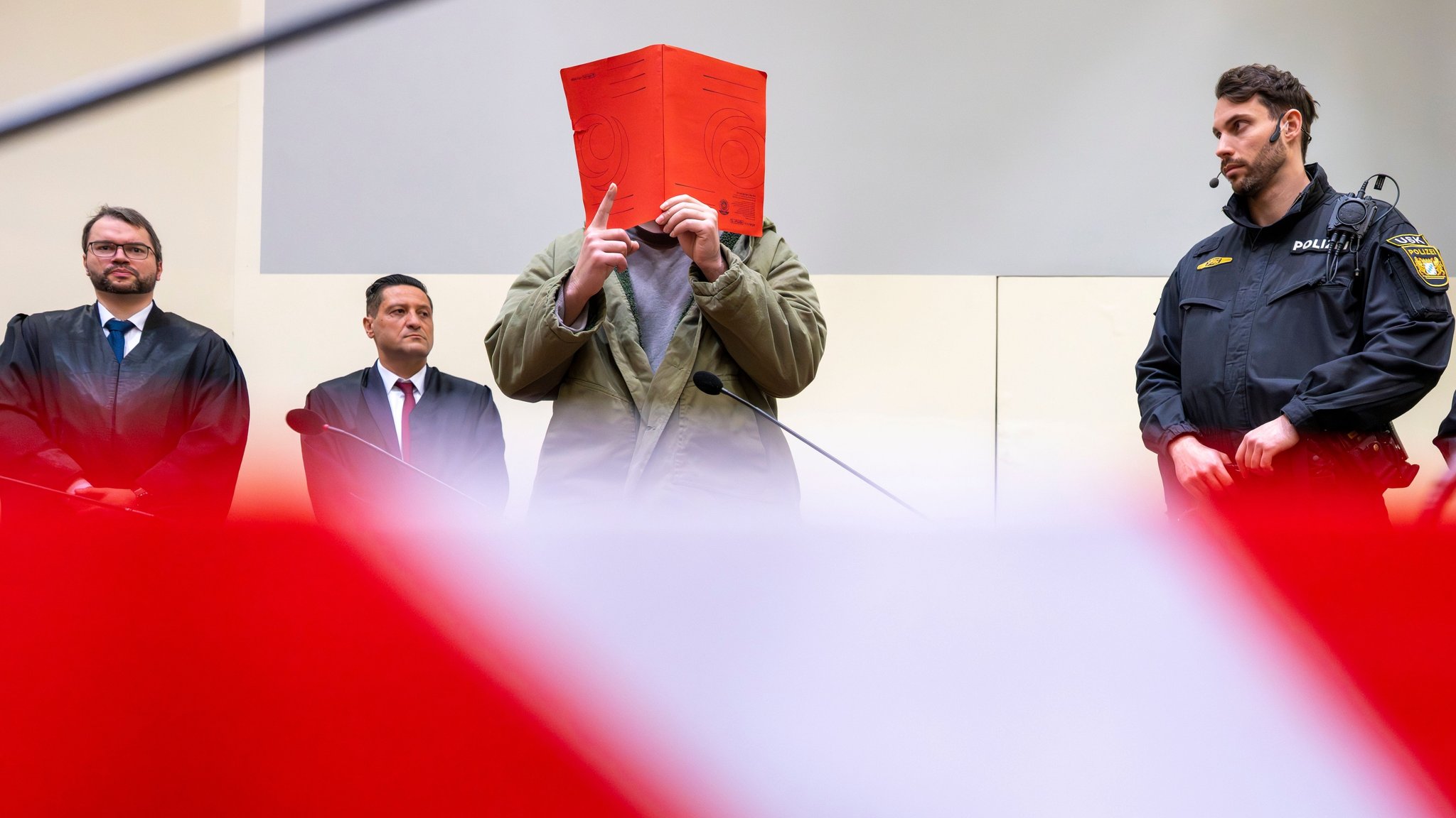 dpatopbilder - 16.01.2026, Bayern, München: Der Angeklagte Farhad N. steht zu Prozessbeginn mit erhobenem rechten Zeigefinger zwischen seinen Anwälten Johann Bund (l) und Ömer Sahinci (2.v.l.) und Polizeibeamten im Prozesssaal an seinem Platz. Farhad N. ist nach einem Auto-Anschlag am 13. Februar 2025 auf eine Demonstration in München des zweifachen Mordes angeklagt. Foto: Peter Kneffel/dpa +++ dpa-Bildfunk +++