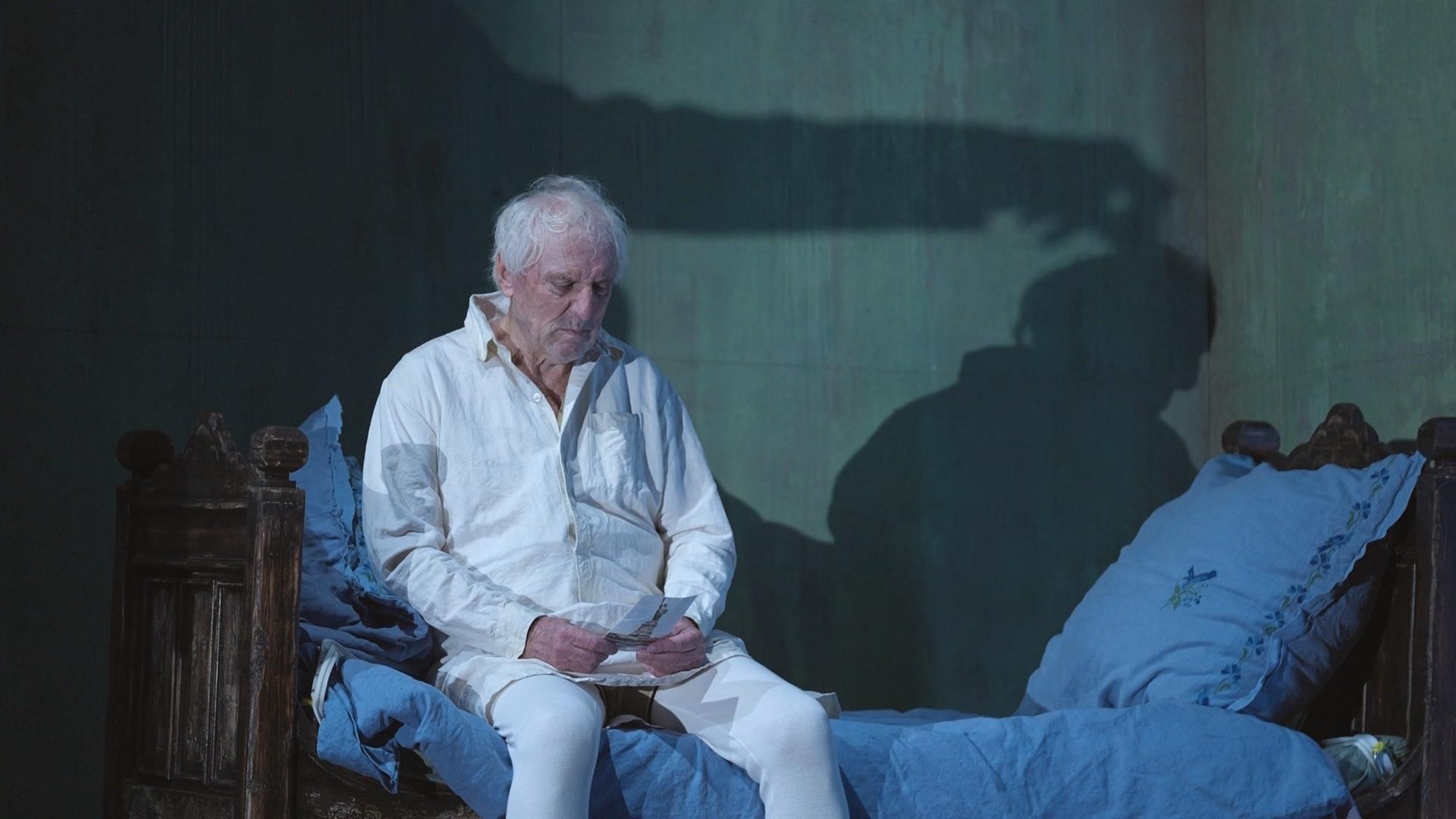 Ein Mann sitzt auf einer Theaterbühne auf einem Bett