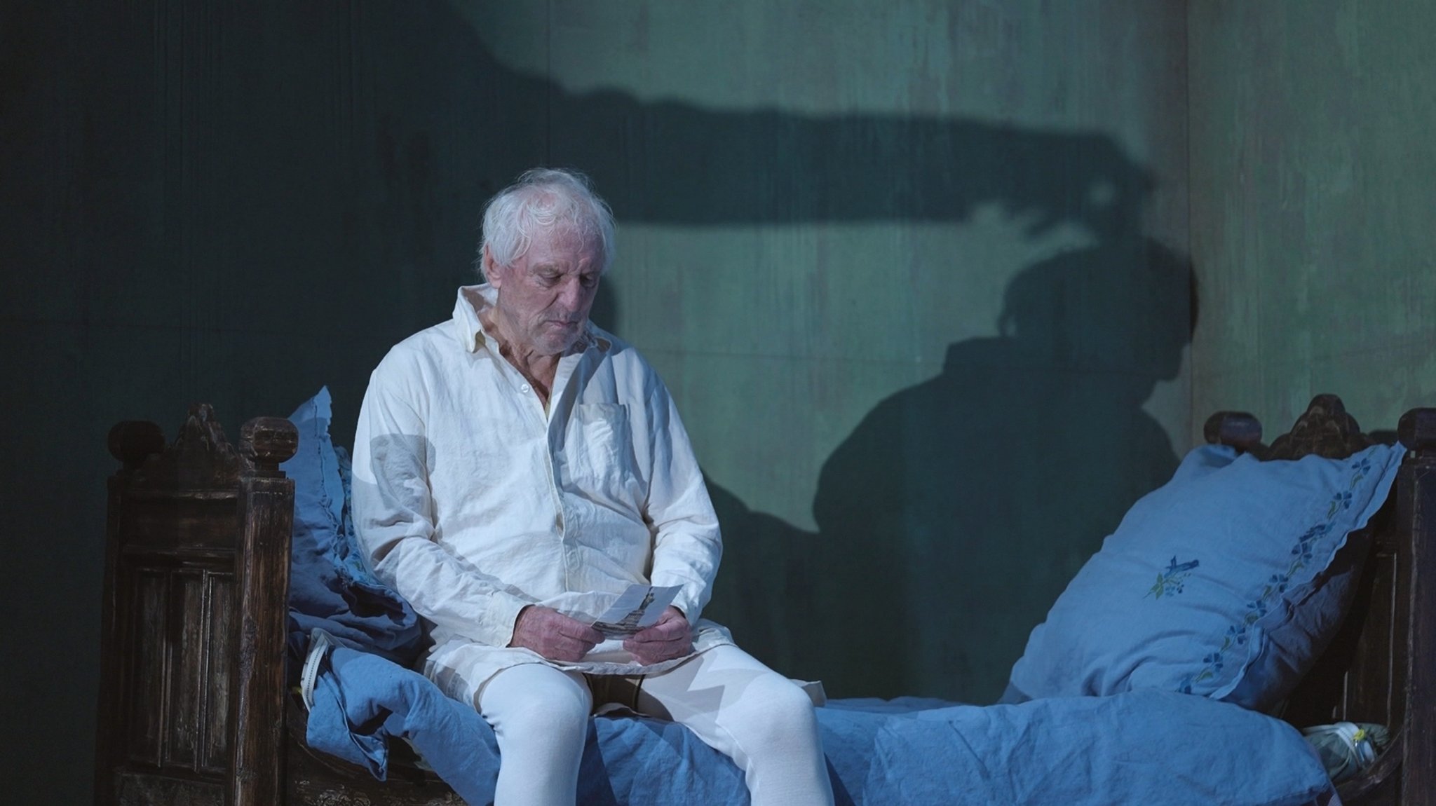 Ein Mann sitzt auf einer Theaterbühne auf einem Bett