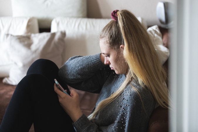 Eine junge Frau sitzt alleine in ihrer Wohnung und hält über ihr Smartphone Kontakt zu ihren Freunden und ihrer Familie.