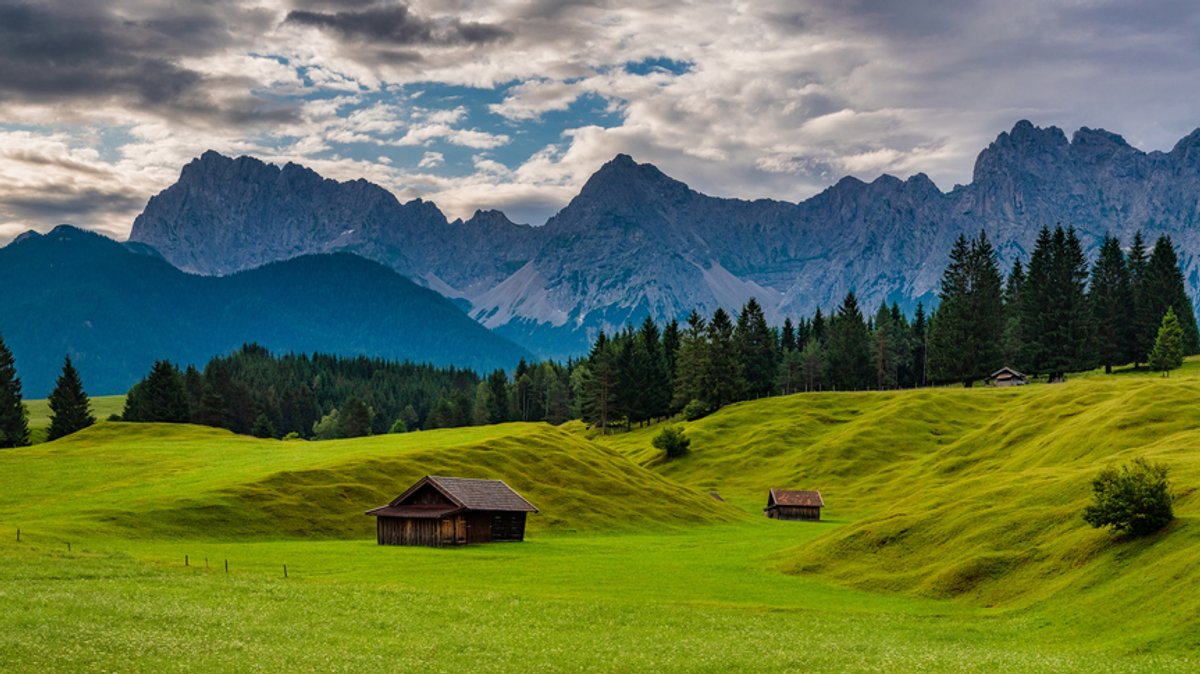 Grüne Buckelwiesen mit drei kleinen Hütten,  vorn im Hintergrund die grauen Felsgipfel des Karwendels bei Mittenwald, darüber leicht bewölkter Himmel 