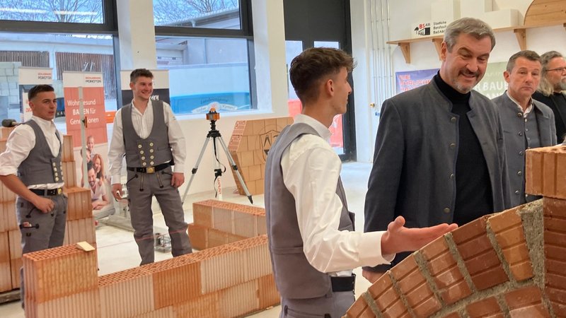 Mit Unterstützung der Berufsschüler darf auch Ministerpräsident Markus Söder (CSU) einen Ziegel setzen. | Bild: BR / Jonas Breitner Mit Unterstützung der Berufsschüler darf auch Ministerpräsident Markus Söder (CSU) einen Ziegel setzen.