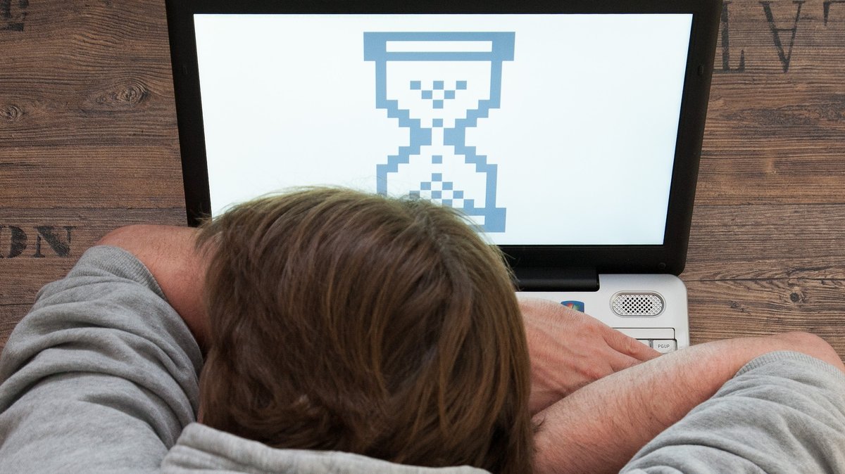 Ein Mann liegt mit seinem Kopf auf der Tastatur eines Laptops, auf dem eine Sanduhr gezeigt wird.