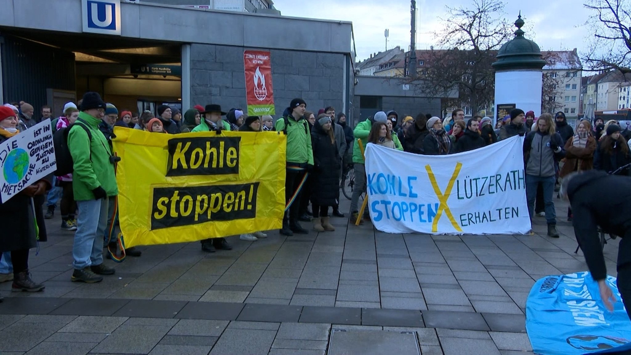 Demonstrierende stehen mit bunten Bannern in der Hand vor einem U-Bahnzugang.