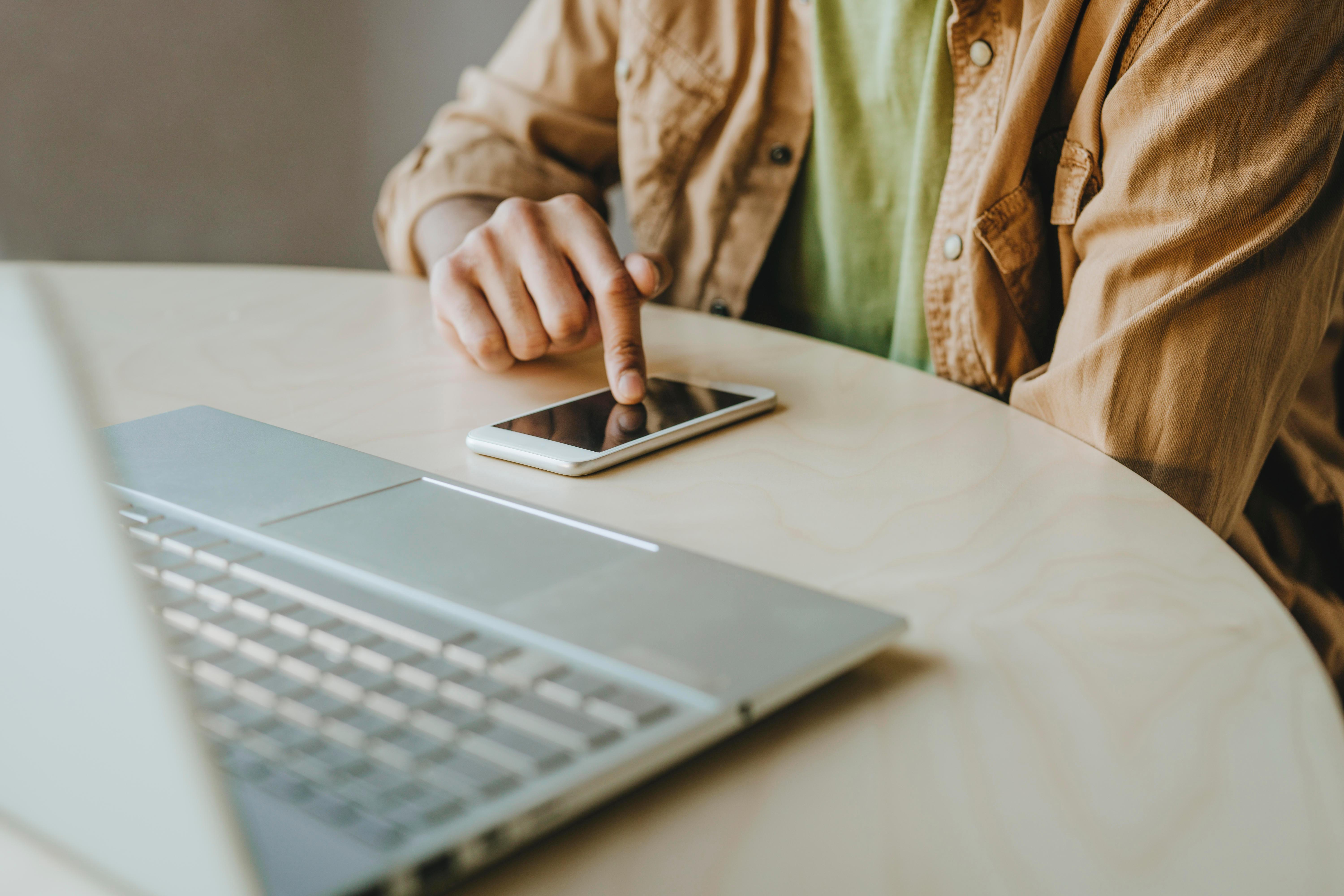 Mann tippt auf sein Smartphone, daneben steht ein Laptop