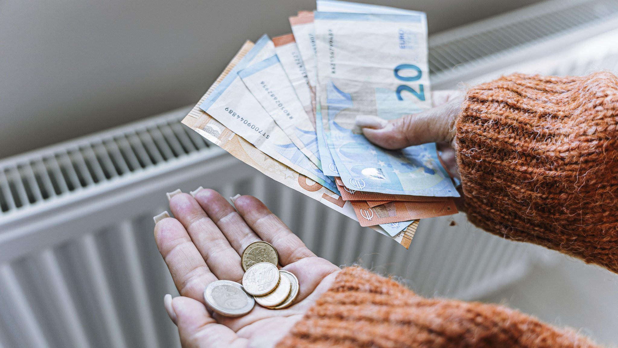 (Symbolbild) Vor einem Heizkörper hält eine Person Geldscheine und Münzen in den Händen.