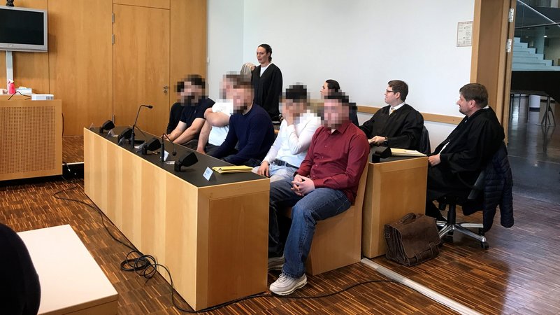 Blick auf die Anklagebank im Augsburger Amtsgericht, auf der fünf junge Männer aufgereiht sitzen. Dahinter ihre Anwälte in schwarzen Roben.