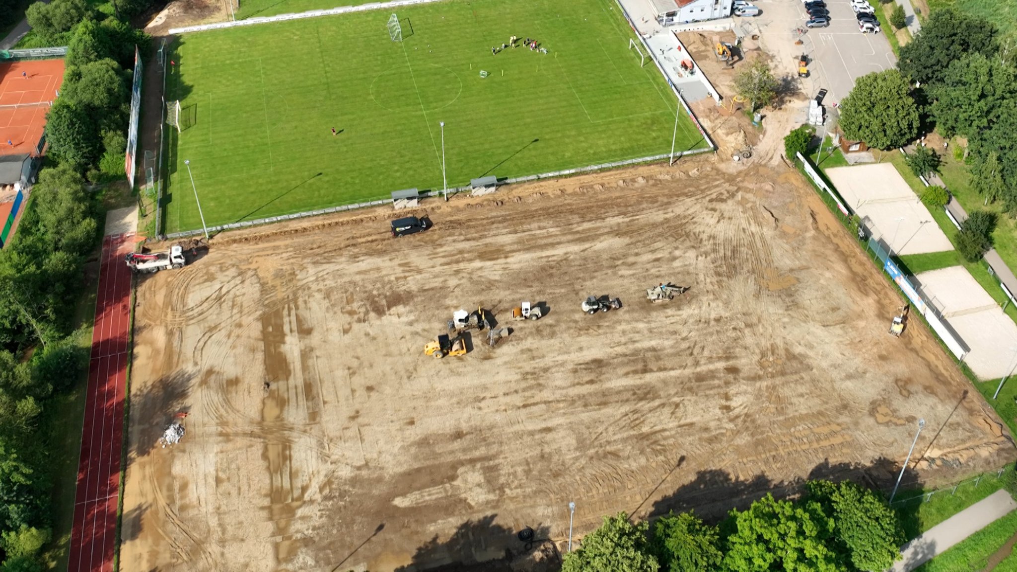 Vogelperspektive: Baumaschinen stehen auf einem Feld ohne Rasen neben einem Fußballfeld mit Rasen.