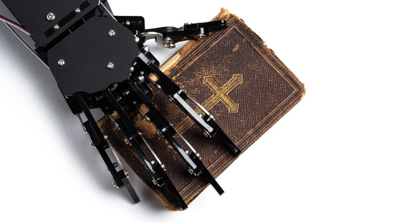 Symbolbild: Hand eines echten Roboters mit einer alten Bibel. | Bild: picture alliance / Zoonar | Anatolijs Jascuks Symbolbild: Hand eines echten Roboters mit einer alten Bibel.