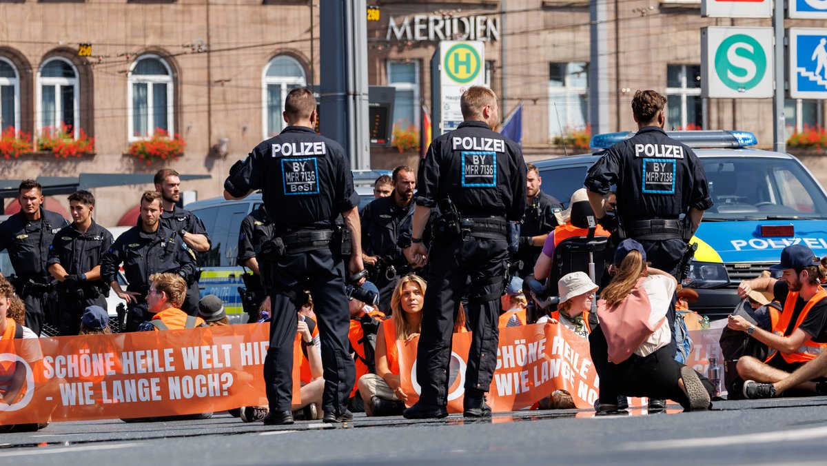 Mehrere Frauen und Männer der Gruppe "Letzte Generation" bei einer Straßenblockade am Nürnberger Hauptbahnhof. Auf einem Banner steht: Schöne heile Welt hier - Wie lange noch?