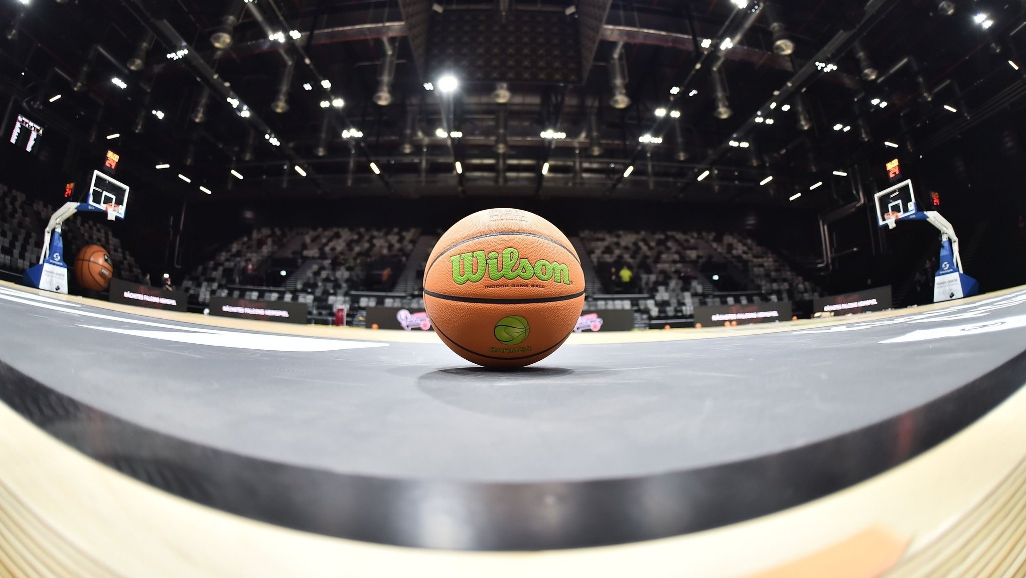 Ein Basketball liegt auf einem Spielfeld in einer Halle, im Hintergrund sind Ränge mit Zuschauern zu sehen.