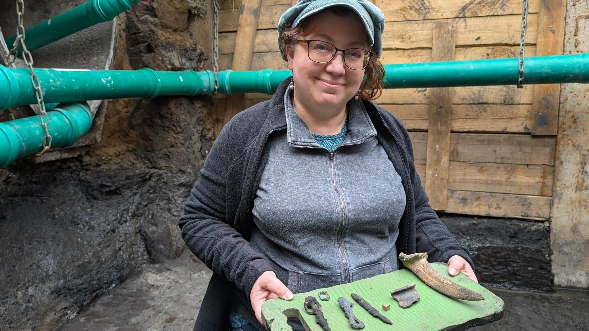 Eine Frau lächelt in die Kamera. Sie hält ein grünes Brett, darauf liegen Gegenstände, die bei einer Ausgrabung gefunden worden. 