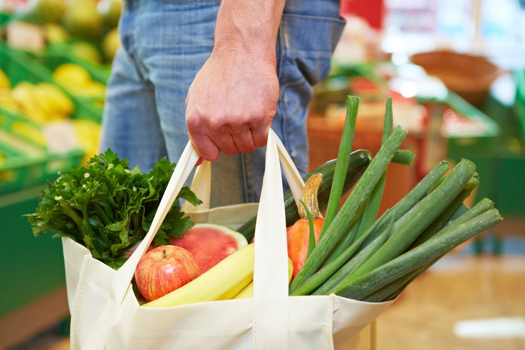 Ein Mensch trägt eine Einkaufstasche, die mit Obst und Gemüse gefüllt ist.