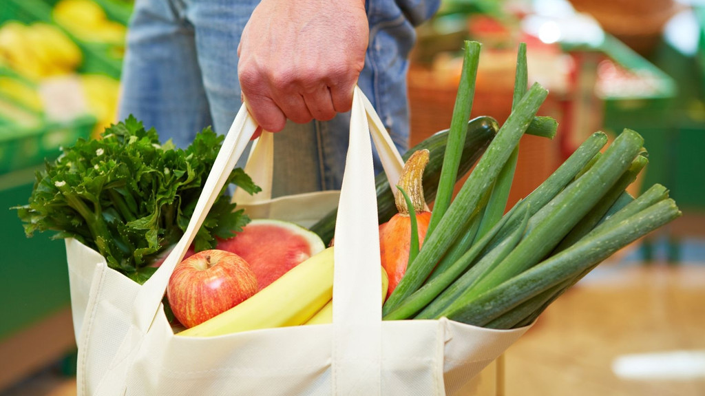 Ein Mensch trägt eine Einkaufstasche, die mit Obst und Gemüse gefüllt ist.