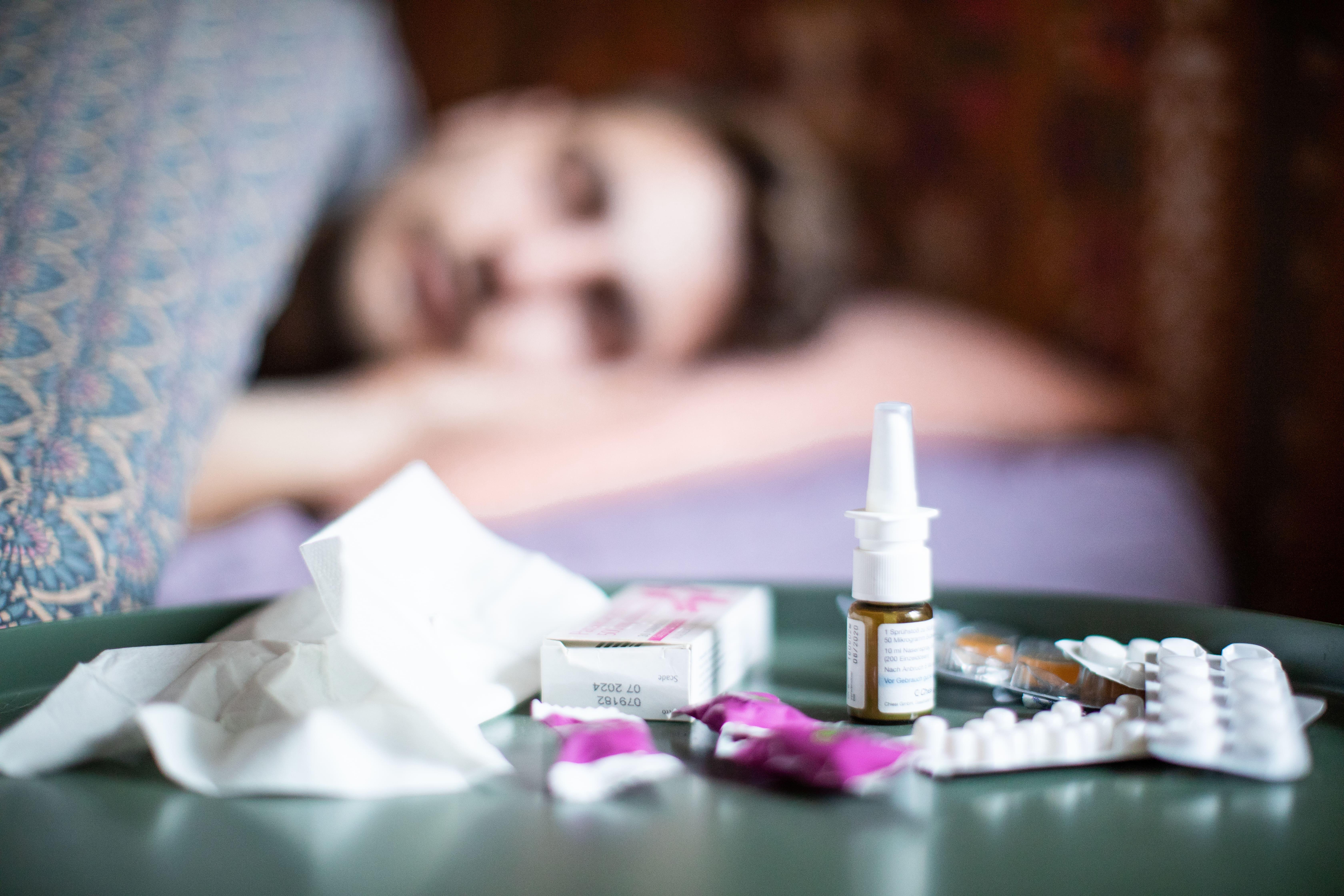 Eine Person liegt krank im Bett, im Vordergrund Medizin.