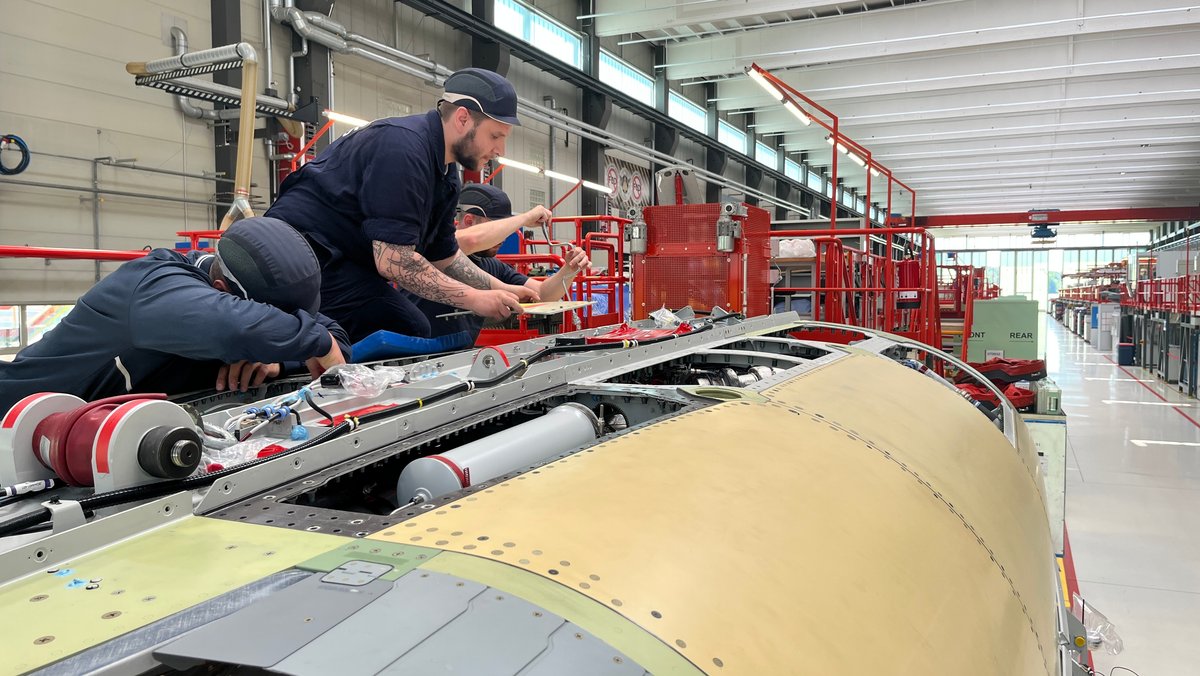 Drei Männer schrauben an einem Flugzeugteil. Dahinter sieht man die Halle, in der die neuen Quadriga-Eurofighter endgefertigt werden.
