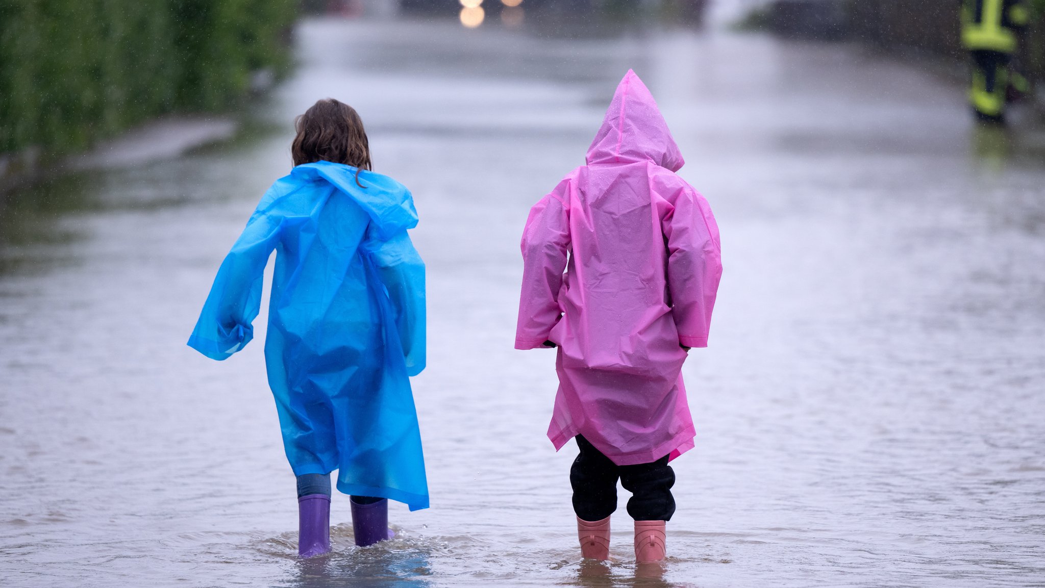 (Symbolbild) Zwei Kinder in Regenmäntel waten durch knöchelhohes Wasser. Auch am Mittwoch fällt in einigen bayerischen Regierungsbezirken aufgrund des Hochwassers der Präsenzunterricht aus.