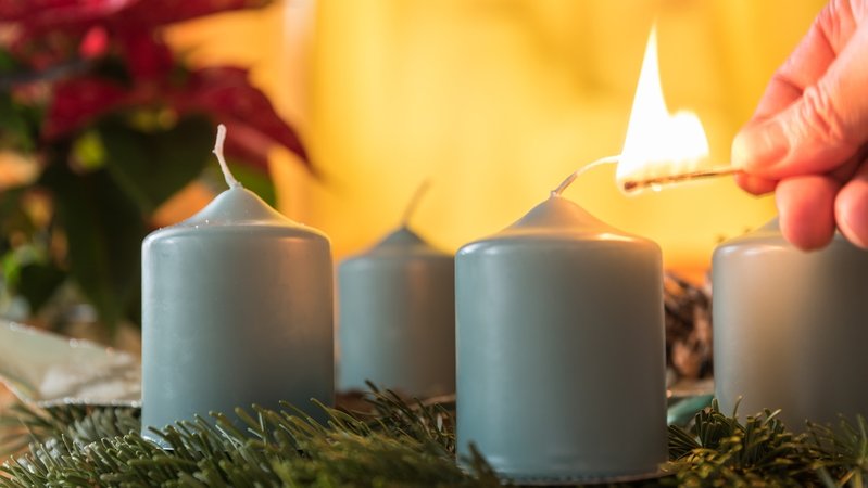 Brennendes Streichholz, mit dem die erste von vier weißen Kerzen auf einem Adventskranz angezündet wird.