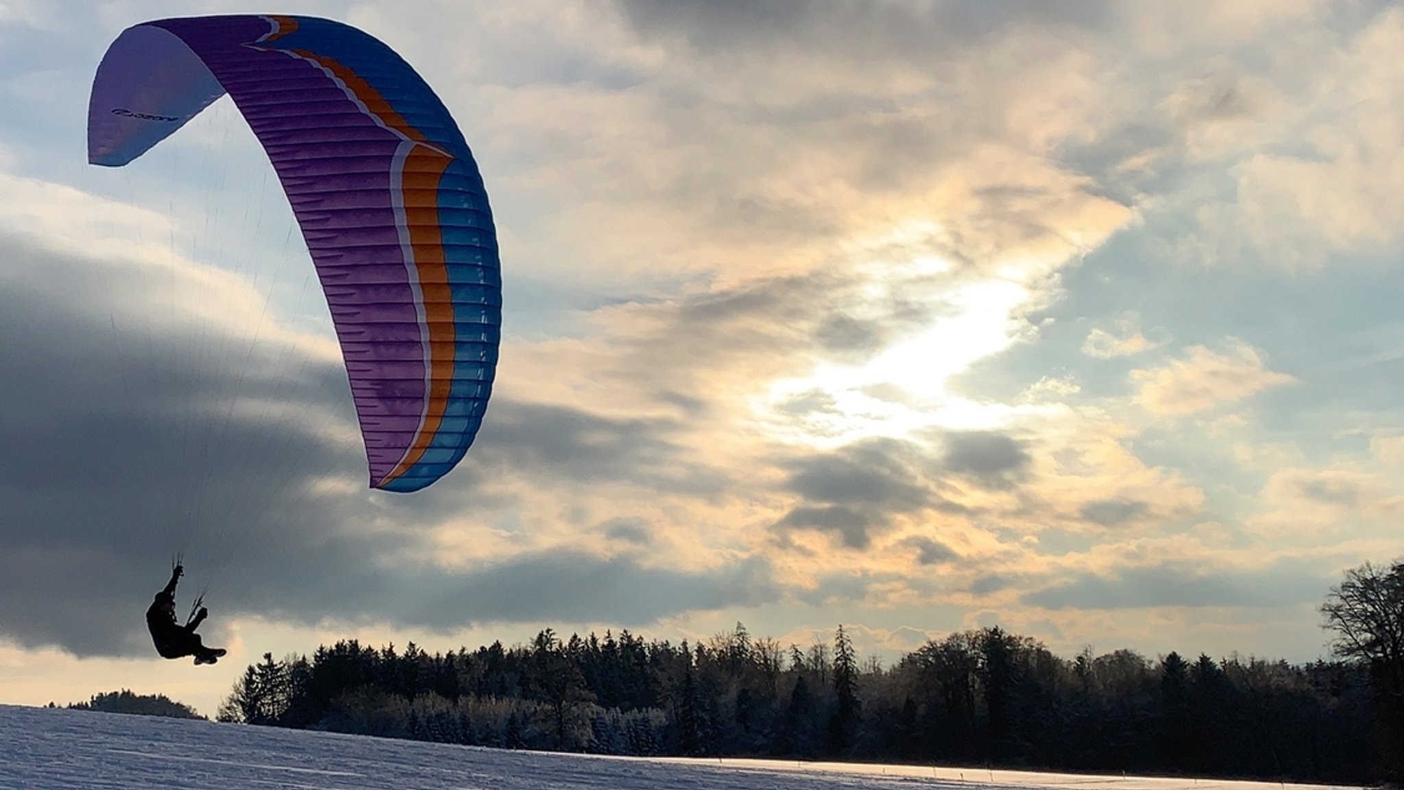 Symbolbild: Ein Gleitschirmflieger über einer schneebedeckten Wiese