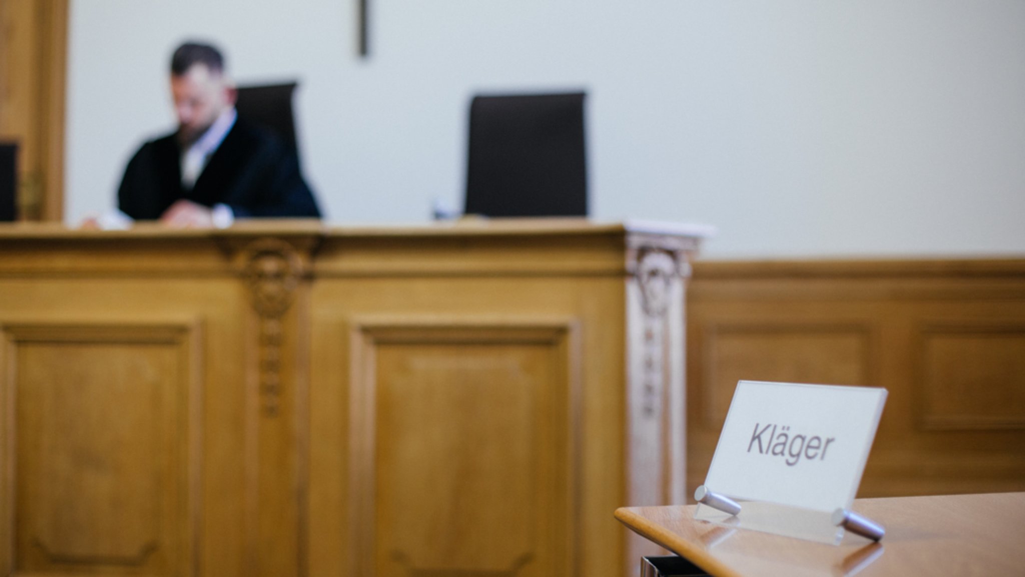 Gerichtssaal. Ein Richter sitzt auf einem Stuhl. Auf einem Tisch davor steht ein Schild "Kläger".