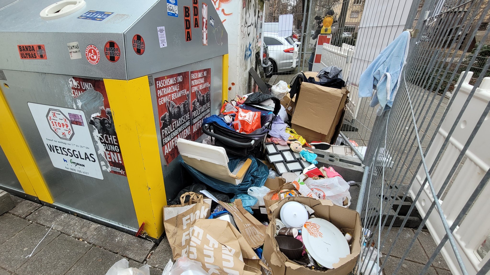 Müll neben einem Altglascontainer. Der Bereich ist mit einem Abuzaun abgesperrt. 