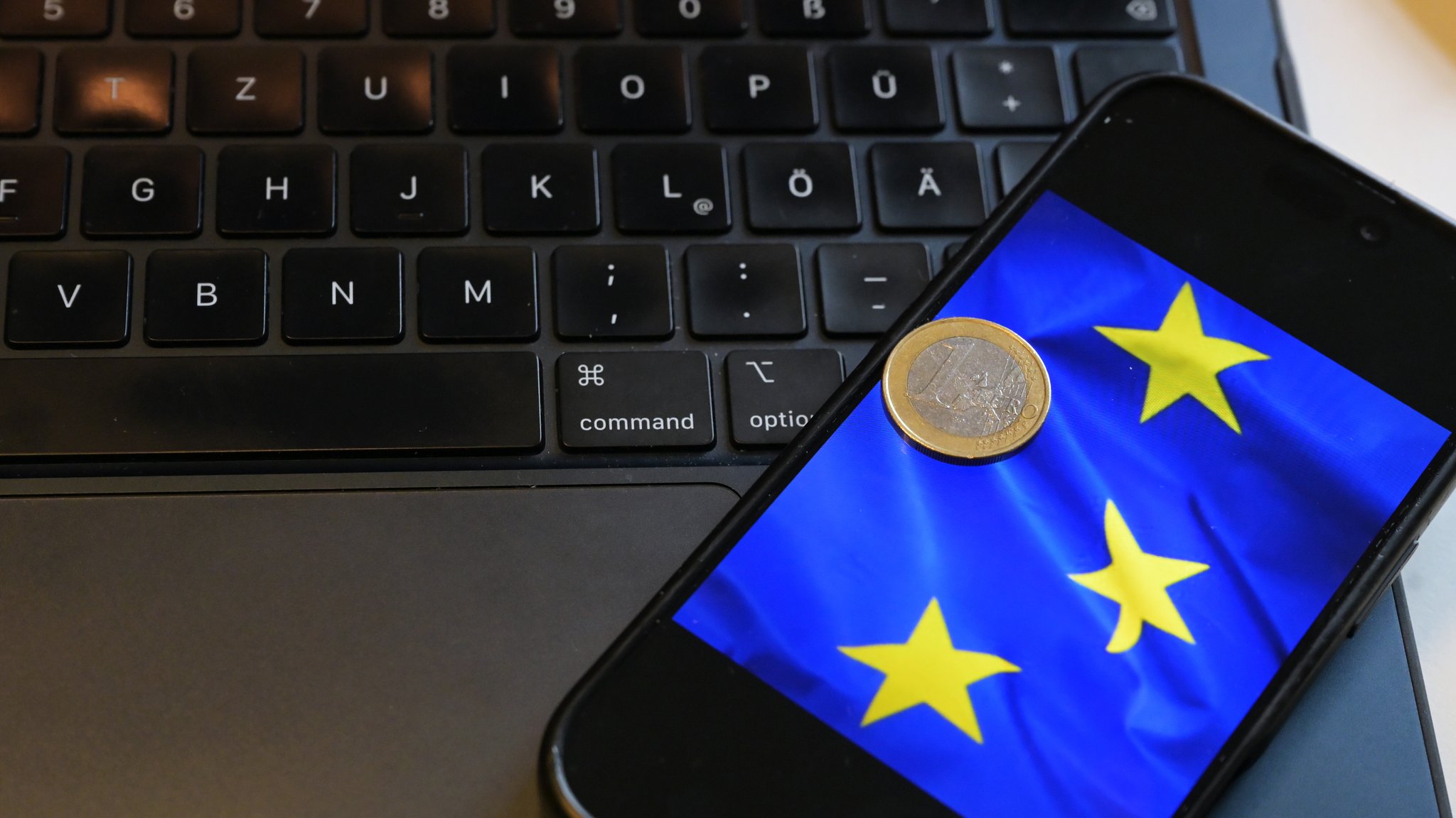 (Symbolbild) Eine Ein-Euro-Münze liegt auf einem Smartphone, das ein Bildausschnitt der Europaflagge zeigt
