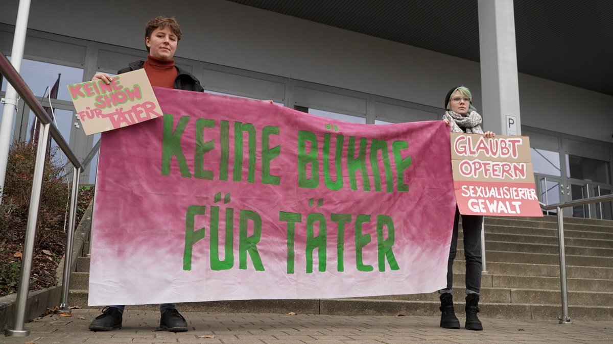 Zwei Frauen stehen mit zwei Plakaten und einem großen Transparent vor der Bamberger Brose Arena. "Keine Show für Täter", "Glabut Opfern sexualisierter Gewalt" und "Keine Bühne für Täter" steht auf ihren Mitbringseln. 
