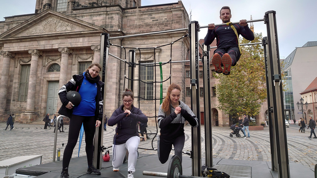 Drei Frauen und ein Mann trainieren mit einem Gewicht und an einer Klimmzugstange draußen im Freien auf dem Jakobsplatz in Nürnberg.