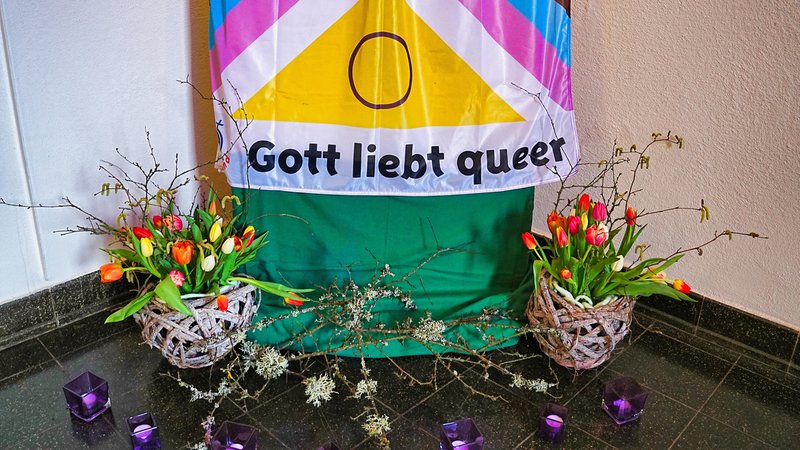 Eine bunte Fahne mit der Aufschrift "Gott liebt queer" schmückt die Ecke eines Raumes. (Symbolbild)