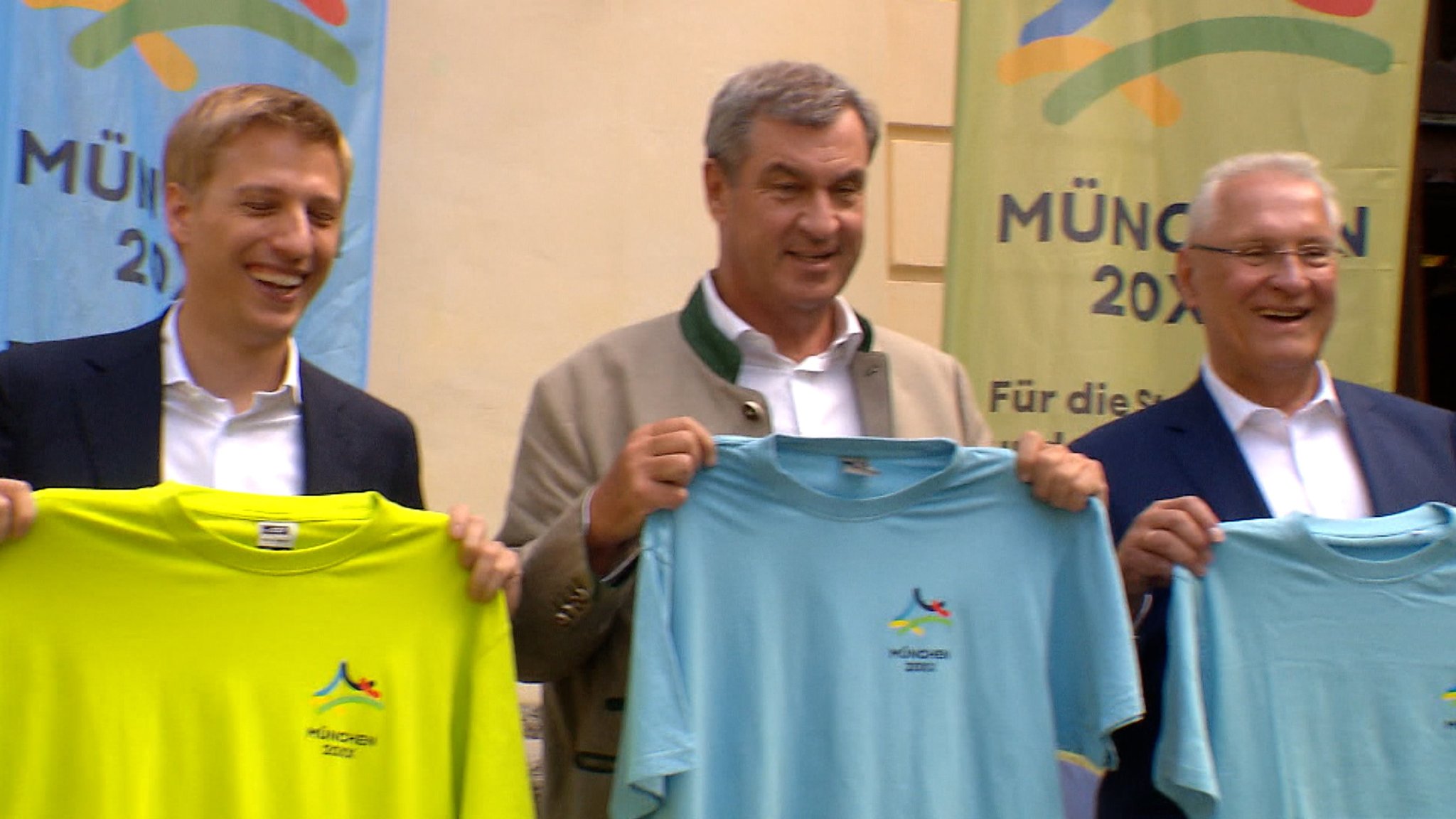 Münchens Oberbürgermeister Krause, Bayerns Ministerpräsident Söder und Innenminister Herrmann halten T-Shirts zur Olympiabewerbung hoch.