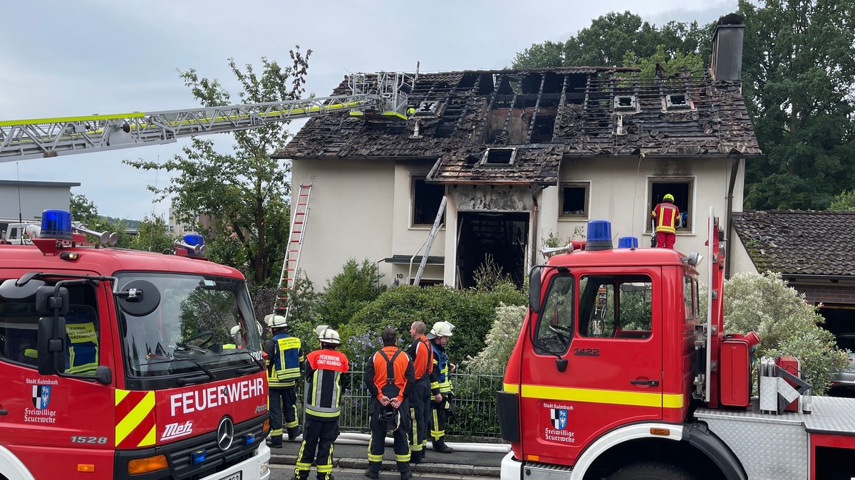 Feuerwehrleute und zwei Einsatzfahrzeuge stehen vor einem Haus, das schwer durch einen Brand gezeichnet ist. 