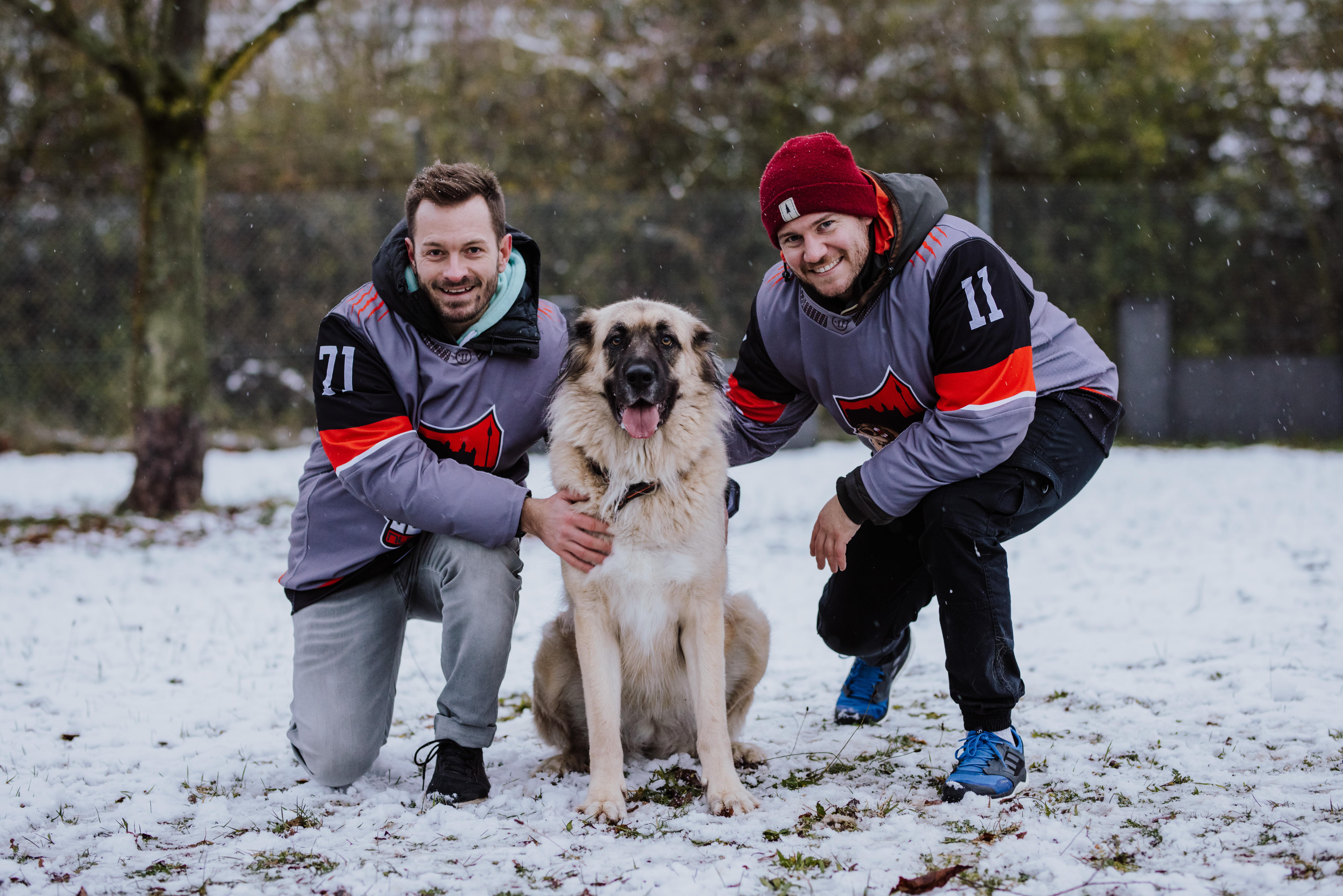 Ein Hund sitzt im Schnee zwischen zwei lächelnden Eishockeyspielern der Nürnberg Bears. 