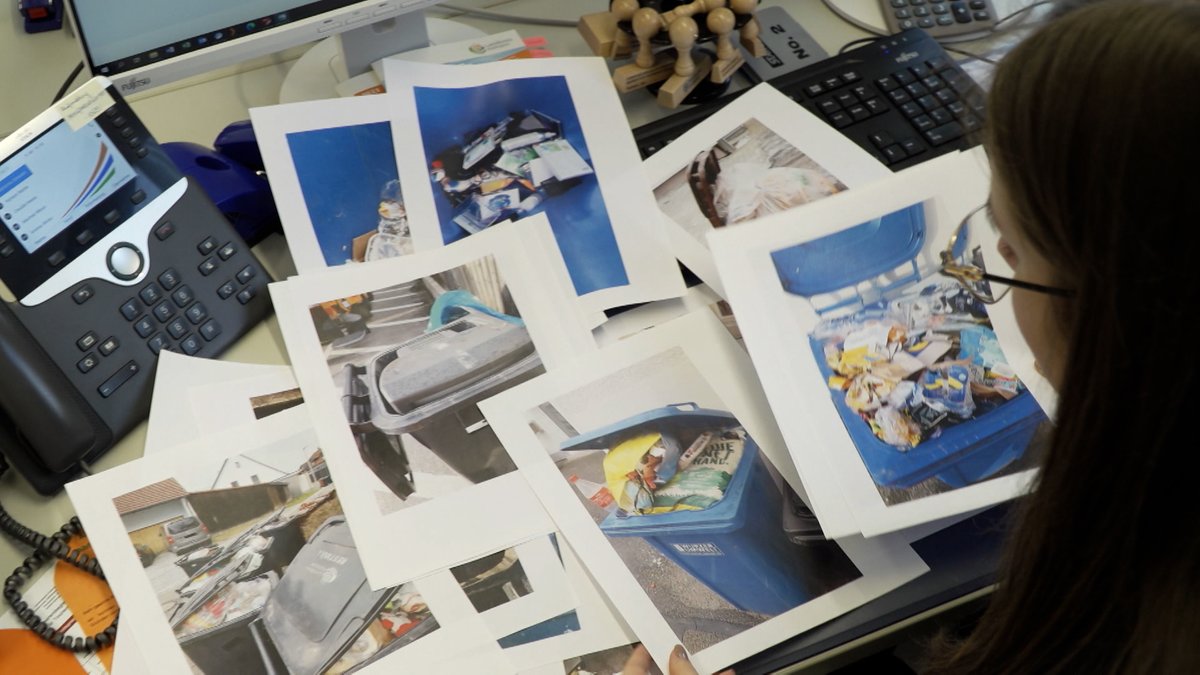 Eine Frau sitzt an einem Schreibtisch, vor ihr sind Fotos von vollen Mülltonnen ausgebreitet.